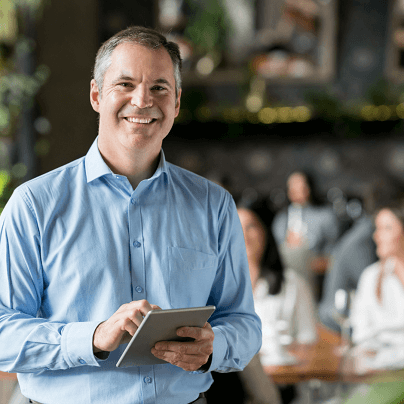 A man wearing a light blue shirt stands smiling while holding a tablet. He is indoors, with a blurred background showing people sitting at tables, suggesting a casual or professional setting like a cafe or meeting space. Perhaps he's reading about self care tips to share with his audience.