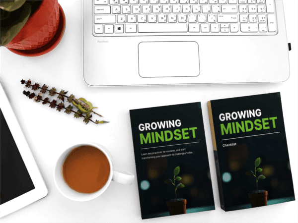 A workspace with a laptop, tablet, coffee cup, and two books titled "Growing Mindset" on a white desk. A small plant in a red pot is nearby, adding a touch of greenery to the scene.